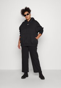 Čierna oversize mikina s predným vreckom a kapucňou so šnúrkou, so malým bielym logom Nike. V páre s čiernymi širokými nohavicami a členkovými čižmami.