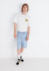 Hvit t-skjorte med "Alfredo's" grafikk, lyseblå denimshorts og svarte joggesko med gule og røde detaljer. Glatte bomull- og denimteksturer.