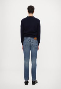 Pull en maille bleu marine avec un col rond, associé à un jean en denim bleu clair avec un patch en cuir et deux poches arrière, porté avec des chaussures noires.