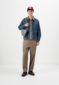 Young man wearing red cap, denim jacket over plaid shirt, beige pants, brown shoes, and carrying a gray crossbody bag on white background.