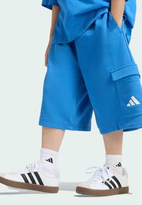 Modré bavlnené šortky so bočnou kapsou a logom, v kombinácii s bielymi teniskami s čiernymi pruhmi a hnedými gumovými podrážkami.