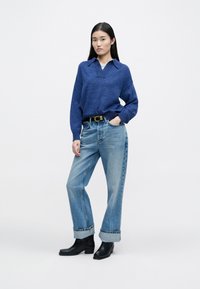 Blauer Strickpullover mit Kragen, kombiniert mit hellblauen hochgeschnittenen Jeans. Als Accessoires werden ein schwarzer Gürtel und Stiefeletten getragen. Einfarbiger Hintergrund.