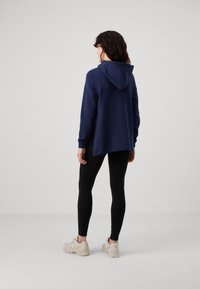 Sudadera con capucha navy de manga larga, con dobladillo redondeado y corte relajado, combinada con leggings negros y zapatillas deportivas de color claro.