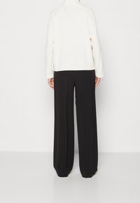 Krämfärgad stickad tröja med hög hals, lös passform och ribbat avslut, kombinerad med högmidjade svarta vida byxor.