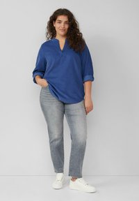 Blaue Bluse mit langen Ärmeln, V-Ausschnitt und aufgerollten Manschetten, kombiniert mit hellgrauen Jeans und weißen Sneakers, auf einem neutralen Hintergrund präsentiert.