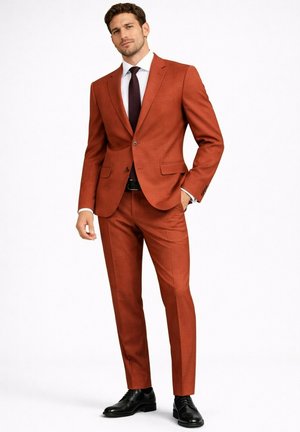 Homme debout avec une main dans la poche portant un costume couleur rouille, une chemise blanche, une cravate sombre, une ceinture noire et des chaussures habillées noires sur fond blanc.