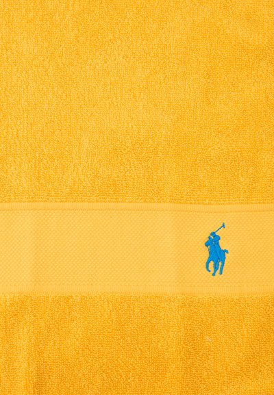 Κίτρινη πετσέτα από περκάλι με υφασμένη ταινία κοντά στο κάτω μέρος, που φέρει ένα μικρό μπλε κεντητό λογότυπο με παίκτη πόλο.