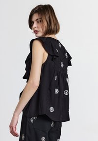 Una giovane donna con capelli corti e castani indossa un top senza maniche nero e pantaloni con motivi floreali bianchi, mentre guarda oltre la spalla contro uno sfondo neutro.