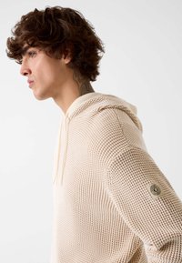 Felpa con cappuccio beige lavorata a maglia con motivo a trama, cappuccio con coulisse e dettaglio logo in metallo sulla manica sinistra. Tessuto morbido e vestibilità rilassata.