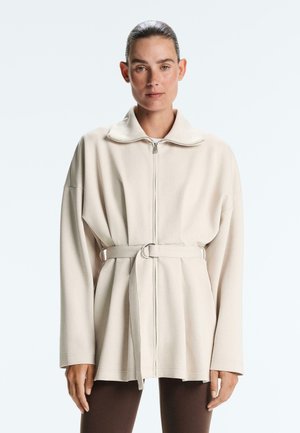 Femeie cu păr dat pe spate, purtând o jachetă bej cu fermoar și curea și pantaloni maro închis, stând pe un fundal simplu și deschis la culoare.