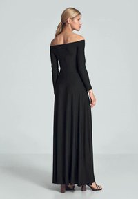 Schwarzes, schulterfreies Kleid mit langen Ärmeln, tailliertem Oberteil und fließendem Rock, aus glattem Stoff hergestellt. Flache Sandalen runden den Look ab.