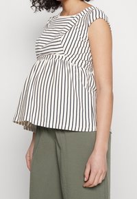 Pruhovaný peplum top v krémové a námořnické modři, s volánkovými detaily a krátkými rukávy, vyrobený z lehkého materiálu. V kombinaci s volnými zelenými kalhotami.