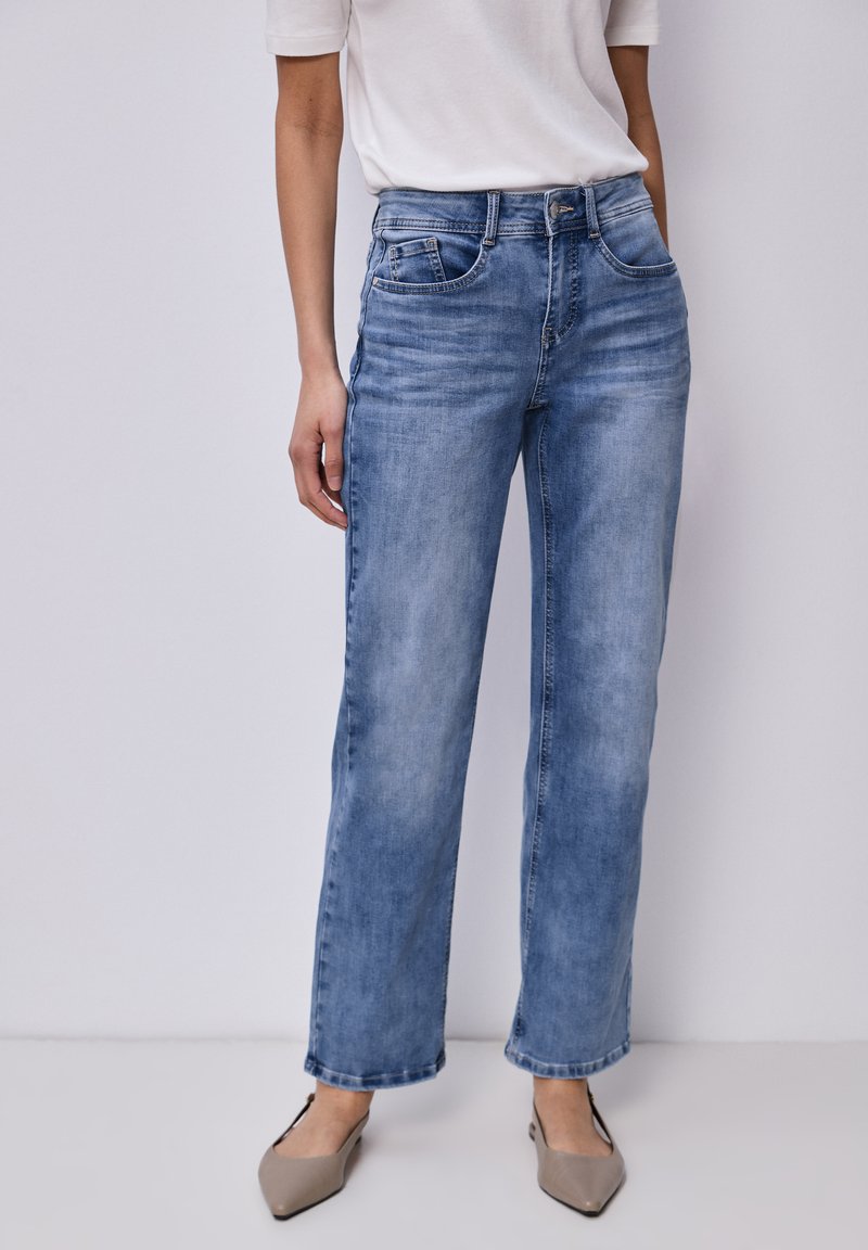 Mujer con jeans de pierna recta azul claro, camiseta blanca de manga corta y zapatos planos beige de punta puntiaguda, de pie contra un fondo liso.