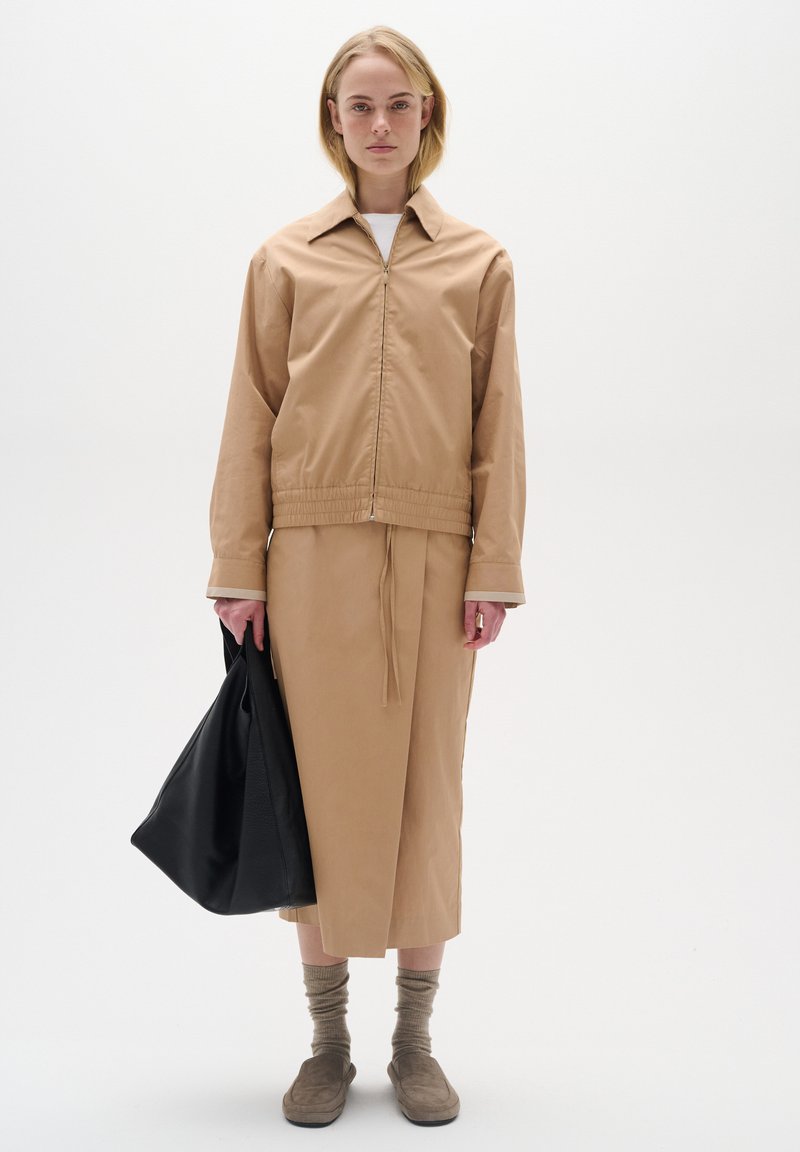 Veste zippée beige avec un col, associée à une jupe assortie, des chaussettes grises et des chaussures à enfiler brunes. Sac fourre-tout noir porté à la main gauche.