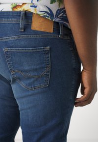 Tummansinisten farkkujen pintakuvioitu viimeistely. Takana tasku, jossa keltaiset ompeleet, sekä nahkainen merkki vyötärönauhassa.