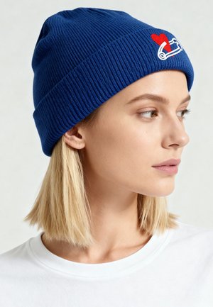 Blue ribbed knit beanie featuring a white safety pin graphic and red heart detail. The beanie is snug-fitting and has a textured appearance.