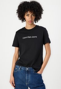Melnā kokvilnas T-kreklā ar īsām piedurknēm un apaļu kakla izgriezumu, uz krūtīm ir balts "Calvin Klein Jeans" logos. Valkāts ar ziliem džinsu biksēm.
