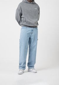 Harmaa vetoketjullinen huppari, jossa on etutasku, yhdistettynä löysiin, vaaleansinisiiin farkkuihin sivutaskulla. Valkoiset tennarit täydentävät asun.