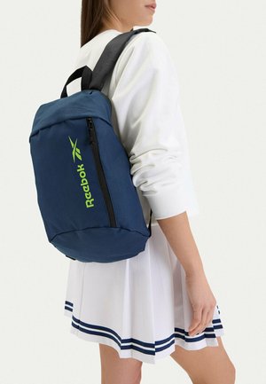 Donna che indossa un top bianco a maniche lunghe e una gonna plissettata con strisce blu navy, che porta una zaino Reebok blu con logo verde neon sulla spalla.
