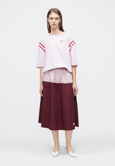 Ροζ μπλουζάκι με κόκκινες ρίγες στα μανίκια, φορεμένο πάνω από μια μπορντό φούστα. Τα παπούτσια είναι άσπρα, γωνιώδη με χαμηλό τακούνι. Απλός σχεδιασμός.