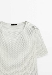 Vit kortärmad stickad t-shirt med ett texturerat, öppet vävmönster och rundad halsringning, som har minimala sömnadsdetaljer.