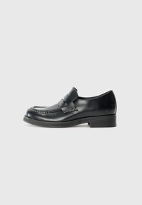 Mocassins en cuir noir avec des coutures de mocassin, une sangle proéminente sur le cou-de-pied, et un petit talon empilé. Fini lisse et bout rond.