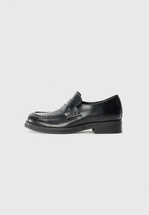 Mocassins en cuir noir avec des coutures de mocassin, une sangle proéminente sur le cou-de-pied, et un petit talon empilé. Fini lisse et bout rond.