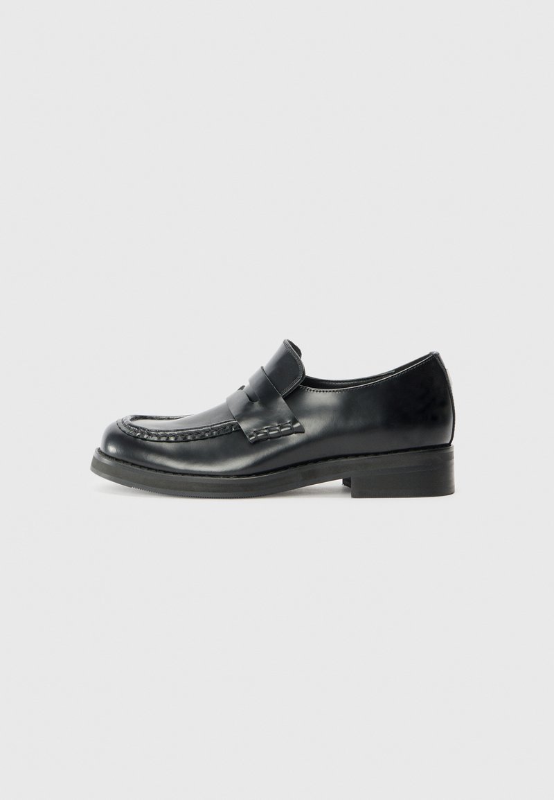 Mocassins en cuir noir avec des coutures de mocassin, une sangle proéminente sur le cou-de-pied, et un petit talon empilé. Fini lisse et bout rond.