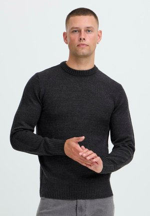 Hombre con cabello corto y pendiente lleva un suéter gris oscuro, de pie contra un fondo claro y liso, con las manos juntas en la cintura.