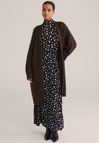 Kobieta ubrana w sukienkę z czarno-białym wzorem w kropki z wysokim kołnierzem, długi ciemnobrązowy kardigan, czarne szpiczaste buty i małe kolczyki koła.