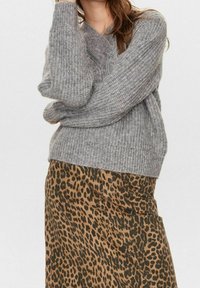 Žena v šedém pleteném svetru s dlouhými rukávy a hnědé sukni s leopardím potiskem stojící před jednoduchým pozadím.