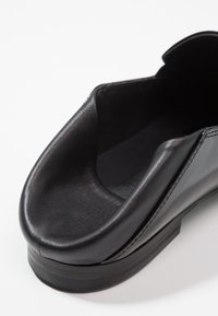 Chaussure en cuir noir avec un bout arrondi, une texture lisse et un design minimaliste. Elle est dotée d'une semelle en caoutchouc basse et d'une large ouverture pour un accès facile.