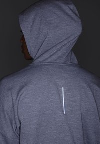 Persoon in een grijze hoodie met een verticale reflecterende strook op de rug, die links draait tegen een donkere achtergrond.