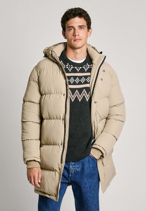 Chaqueta de plumón beige con capucha, que presenta un cierre frontal de cremallera y botones a presión. Llevada sobre un suéter oscuro de punto con un diseño, combinada con jeans azules.