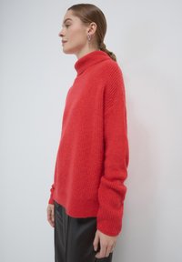 Frau im Profil, die einen leuchtend roten gestrickten Rollkragenpullover und schwarze Hosen trägt, mit zurückgebundenem Haar, vor einer weißen Wand stehend.