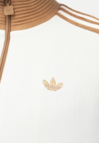 Chaqueta blanca con cremallera que presenta un diseño acanalado y un alto cuello beige. Destacan los acentos en beige con tres franjas y un logo bordado en beige en el pecho.