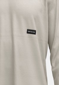 Helles graues Langarmshirt mit strukturiertem Stoff, horizontaler Naht und einem rechteckigen schwarzen Ortovox-Label auf der linken Seite.