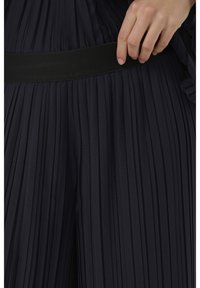 Sötét, pliszírozott anyag, széles fekete derékpánttal; a textúra sima. A közeli nézet kiemeli a finom pliszírozást és a derékpánt részleteit.