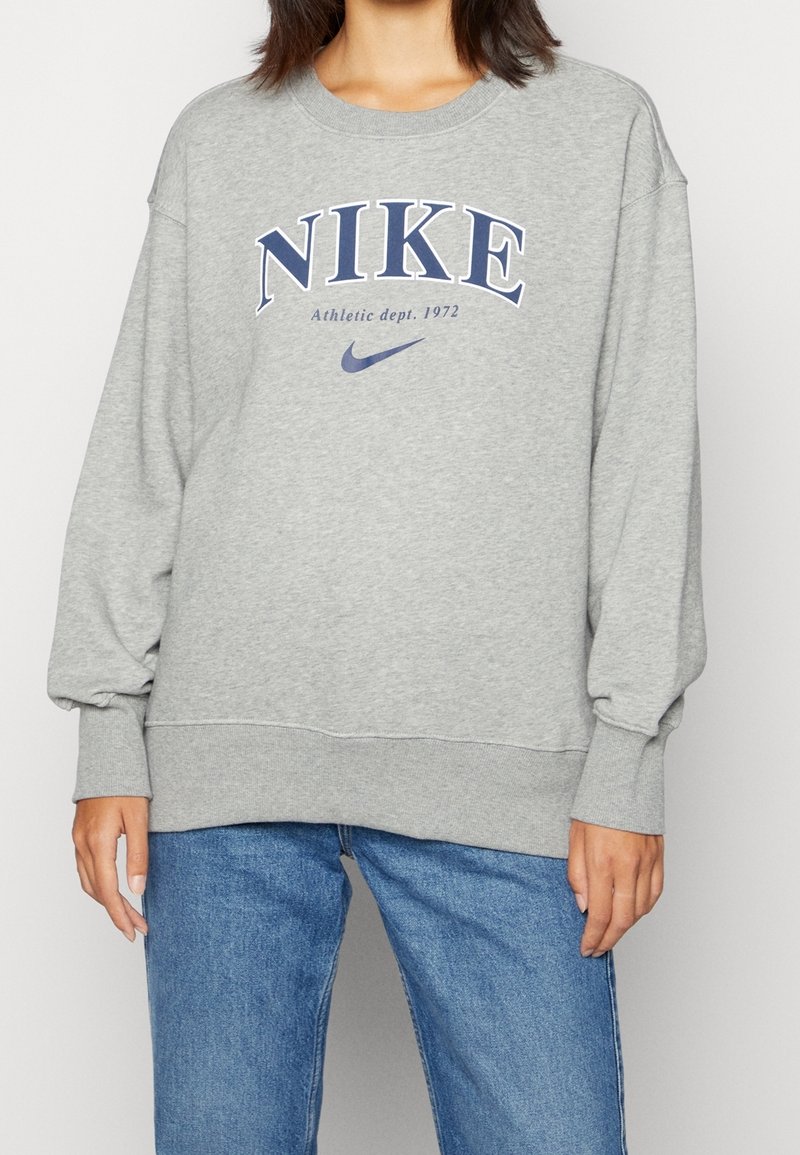 Grå sweatshirt med "NIKE"-logotyp i marinblått, ribbade ärmslut och nederkant, avslappnad passform och mjukt bomullsmaterial. Matt yta.