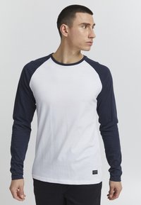 Fehér és sötétkék hosszú ujjú raglan póló, pamutból készült, kerek nyakkivágással és kontrasztos ujjakkal, a szegélyén kis márkacímkével.