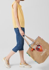 Geel mouwloos topje met kantafwerking, blauwe cropped jeans en beige sneakers; grote bruine tote met een pailletontwerp en kleurrijke pomponnen.