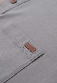 Tela de algodón gris con un bolsillo en el pecho; cuenta con una etiqueta de cuero marrón cosida en el bolsillo; textura suave y diseño simple.