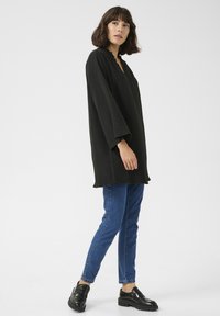 Överstor svart topp med vågig halsringning, långa ärmar, matchad med slim-fit blå jeans och svarta kraftiga loafers. Slät textur.