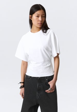 Νεαρή γυναίκα με μακριά σκουρόχρωμα μαλλιά, που φοράει μια φαρδιά λευκή κοντομάνικη μπλούζα και μαύρα ψηλόμεσα τζιν, στέκεται μπροστά σε ένα απλό φόντο.