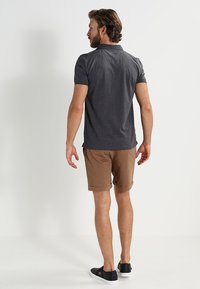 Kullsvart polo skjorte med krage, parret med lysebrune shorts. Modellen har på seg svarte slip-on sko. Glatt stoff med en avslappet passform.