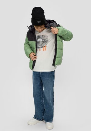 Grüne und schwarze gepolsterte Jacke, graues T-Shirt mit Grafik, blaue Jeans, weiße Sneakers und schwarze Mütze mit Logo.
