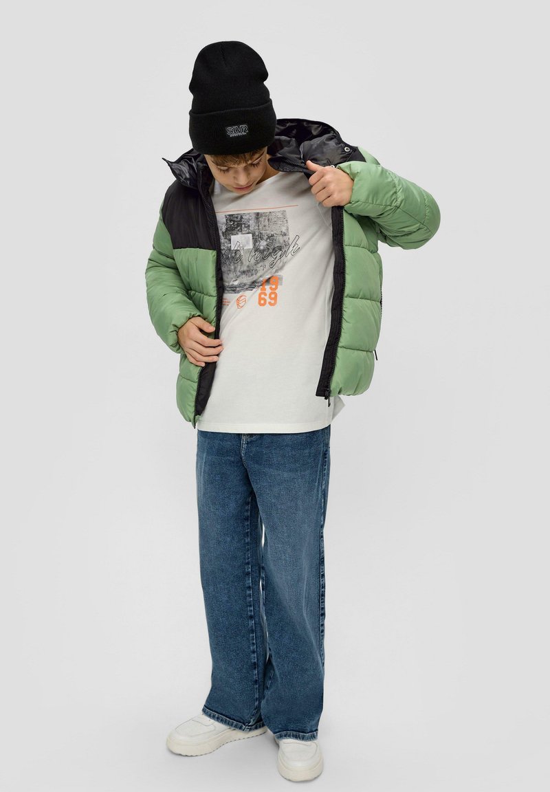 Padded jacket w kolorze zielonym i czarnym, szara koszulka graficzna, niebieskie dżinsy, białe trampki oraz czarna czapka beanie z logo.