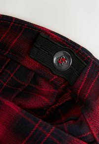 Červená a černá kostkovaná tkanina s texturovaným povrchem, obsahující černý pas, zapínání na knoflík a viditelné detaily šití.