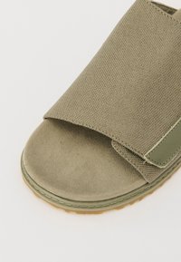 Tongs de couleur vert olive avec une tige en tissu texturé, un intérieur en suède doux et une semelle en caoutchouc. Présente un design de lanière large et superposée.