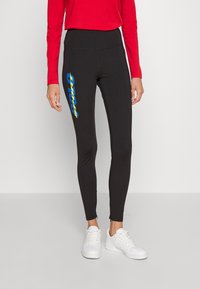 Svarta leggings i stretchtyg med hög midja och en blå- och gulllogotypgrafik på vänster ben. Vita sneakers.